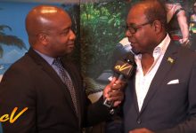 Two men in suits, one holding a microphone, in front of a beach backdrop; interview with Jamaica's Tourism Minister Edmund Bartlett